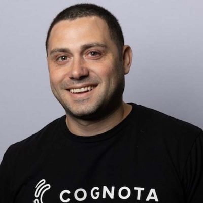 Ryan Austin A smiling man with short dark hair wearing a black "COGNOTA" T-shirt stands against a plain light gray background, ready to enjoy the I4PL after party. - Cognota