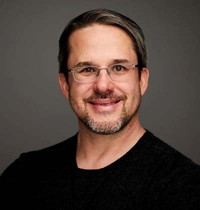 Sabino Marquez, a man with short brown hair, glasses, and a trimmed beard, smiles at the camera. He is wearing a black shirt and is posed against a neutral gray background. - Cognota